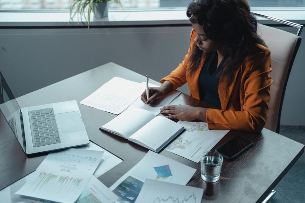 Businesswoman in office writing notes with documents and laptop, focused on analysis and productivity.